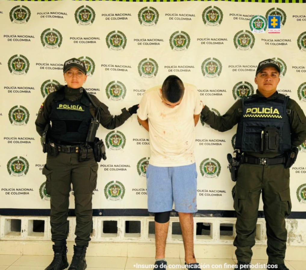 El capturado se encuentra de frente y custodiado por dos uniformados de la Policía Nacional. En la foto se ve de fondo un backing de la Policía Nacional.
