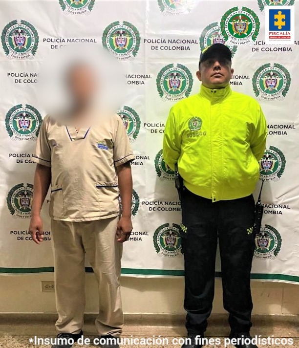 El capturado tiene puesto un uniforme de enfermero, estpa custodiado por un uniformado de la Policía nacional y detrás de ellos, hay un pendón institucional.