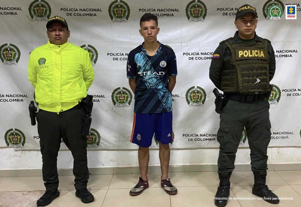 El capturado aparece esposado con sus manos a la espalda, viste pantaloneta y camiseta deportivos y está custodiado por dos uniformados de la policía nacional.