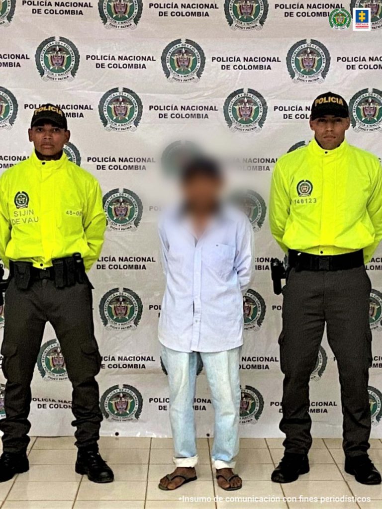 El capturado está esposado y protegido por dos unidades policiales y detrás de ellos está un pendón institucional.