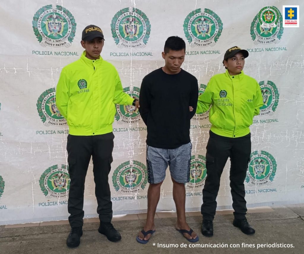 El capturado está con su rostro agachado, esposado a su espalda y custodiado por dos uniformados de la policía nacional.