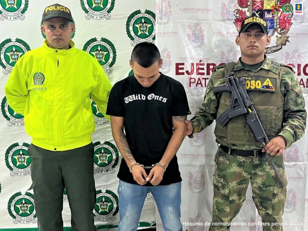 El capturado aparece esposado y con sus manos al frente, está custodiado por dos uniformados de la policía nacional y del ejército.