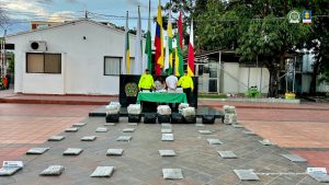 Los dos capturados se encuentran de espaldas y custodiados por dos uniformados de la Policía Nacional. Detrás de la foto hay unas banderas y se observa una mesa verde con el material que se les incautó y en el piso también se observa el estupefaciente.