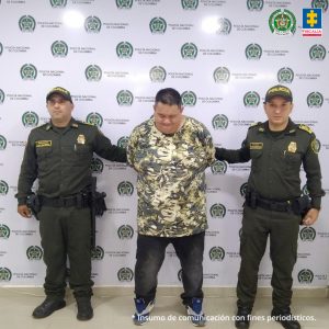 en la imagen se ve una persona detenida bajo custodia de dos integrantes de la Policía Nacional. Detrás suyo un backing institucional.