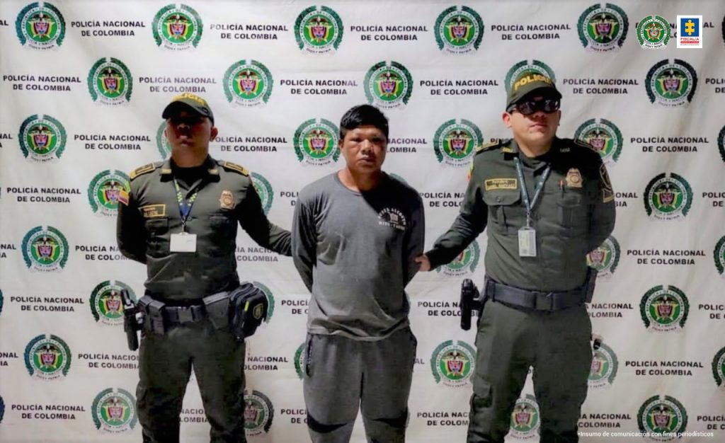 El capturado está esposado con sus manos a la espalda, y tomado de sus brazos por dos uniformados de la policía nacional.