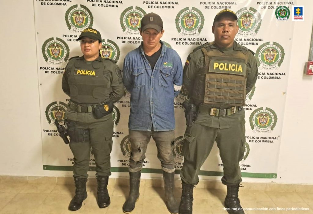 El capturado aparece esposado de manos a su espalda y está custodiado por dos miembros de la Policía Nacional.