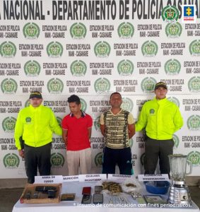 Los capturados están esposados y custodiados por uniformados de la fuerza pública, y los elementos materiales probatorios incautados, están exhibidos en una mesa., con sus manos esposadas, y en una mesa todos los elementos materiales probatorios incautados durante las diligencias de allanamiento.