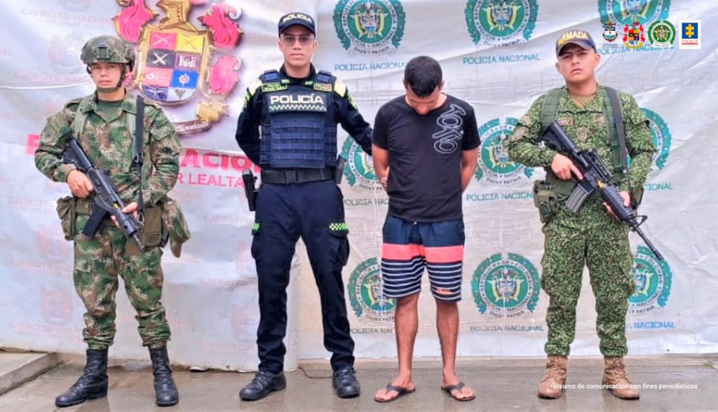 El capturado está vestido con una camiseta negra, pantaloneta azul con rayas rojas y está custodiado por uniformados de la armada nacional, ejército y policía nacional.