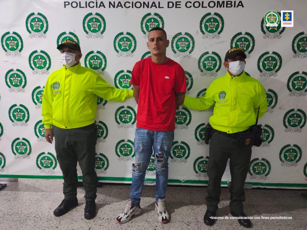 Andrés Felipe Pérez Rubio alias Riosucio, tiene sus manos atras con esposas y rodeado de dos uniformados de la policía nacional, y atrás el pendón de la institución.