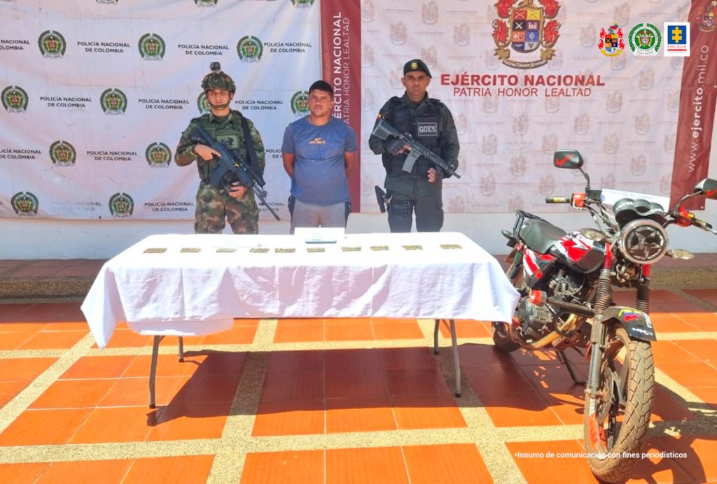 El capturado está esposado, con sus manos a la espalda y está custodiado por un miembro del Ejército Nacional y uno de la Policía, y en una mesa frente a ellos están expuestos los proyectiles incautados.