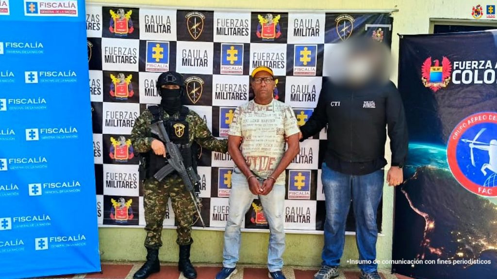 El procesado aparece de frente a la cámara, esposado con sus manos adelante, custodiado por un servidor del CTI Fiscalía y un uniformado del Gaula Ejército. Detrás de ellos, hay varios pendones institucionales.