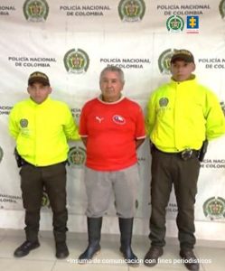 El procesado aparece vestido con una camiseta roja y un pantalón gris. De fondo está el backing de la Policía y dos uniformados.