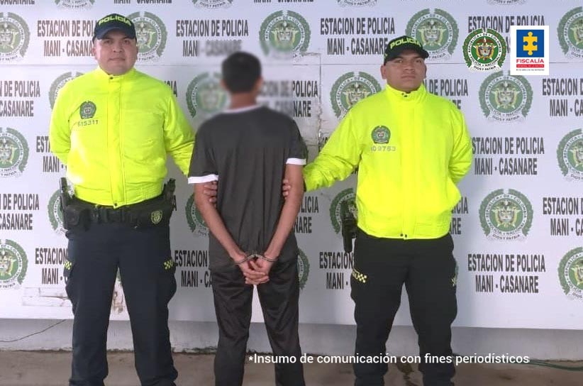 El capturado está de espalda, con sus manos esposadas y dos policías sujetándolo, de fondo hay un pendón institucional