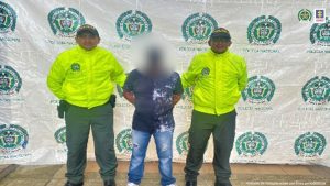 El capturado tiene gorra negra y con su cara agachada, esposado con sus manos a la espalda y custodiado por dos policías.
