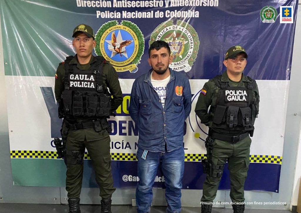 El capturado está esposado con sus manos a la espalda, y está custodiado por uniformados del Gaula de la Policía nacional.