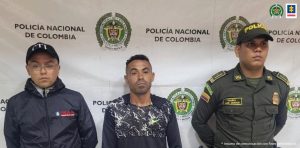 El capturado aparece de pie en medio de dos uniformnados de la policía nacional, fue capturado por ser el presunto delito de violencia intrafamiliar agravada, detrás de ellos hay un pendón institucional.