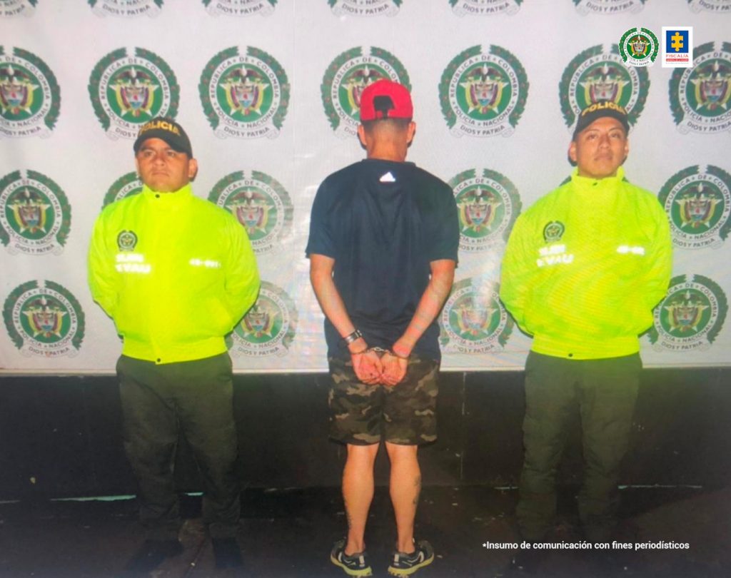 se oberva a un hombre espaldas con gorra roja en una pantaloneta camuflada acompañado de dos uniformados de la policía Nacional