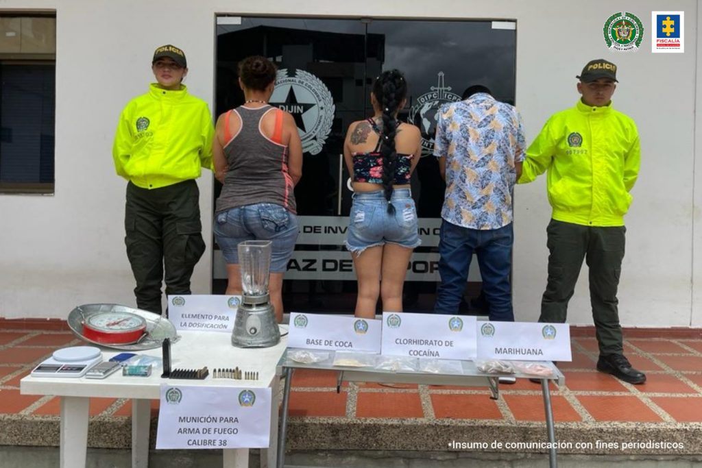 Los tres capturados están de espalda a la cámara, esposados y custodiados por uniformados de la policía nacional, y en una mesa, están los elementos incautados.