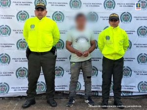 El capturado está custodiado por uniformados de la Policía Nacional, con sus manos esposadas y de fondo un pendón de la policía.