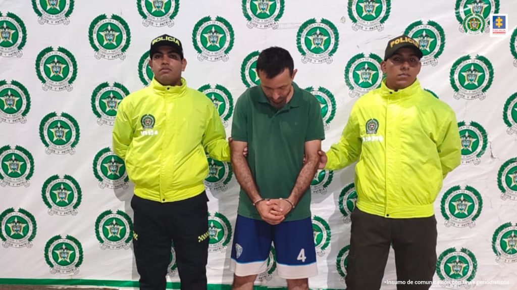 El capturado está en medio de dos uniformados de la policía, y detrás de ellos hay un pendón institucional.