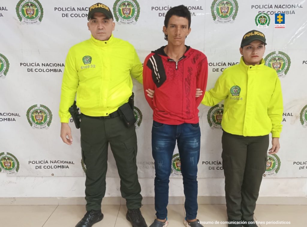 El capturado viste una camiseta roja, está sujetado de los brazos por dos uniformados de la policía nacional y atrás de ellos un pendón institucional.