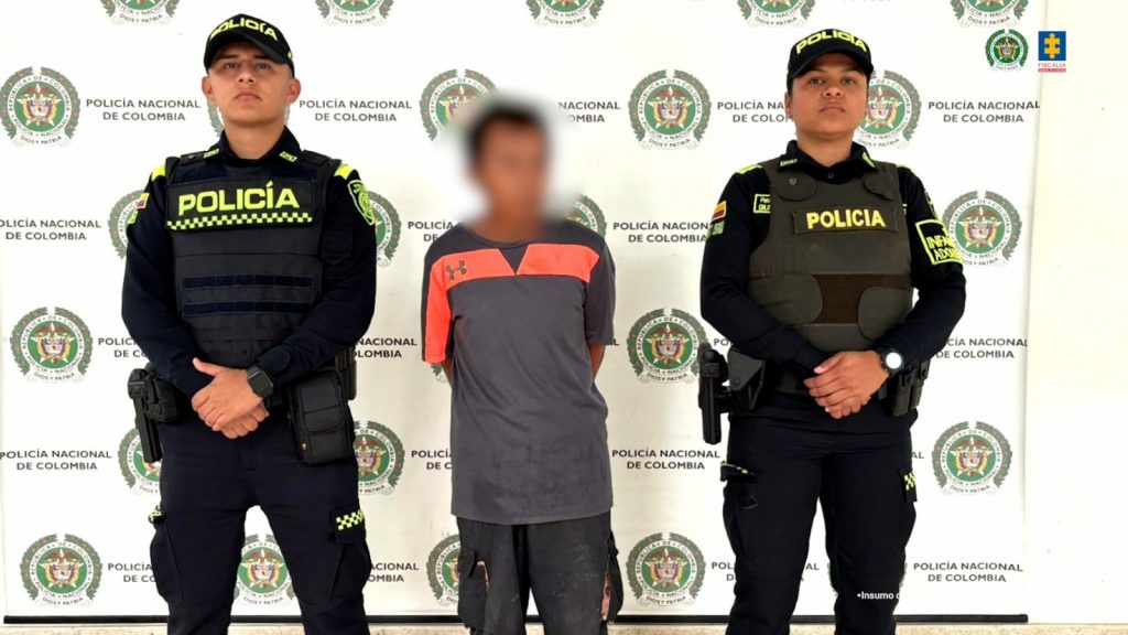 El capturado está custodiado por dos uniformados de la Policía, con sus manos esposadas a la espalda y de fondo un pendón institucional.