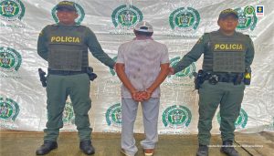 El capturado está esposado con sus manso a la espalda y custodiado por dos hombres de la policia nacional.