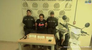 En la imagen aparece la mujer aparece con sus manos a la espalda, custodiada por dos uniformados dela Policía, y en una mesa frente a ellos, un revólver, munición y un equipo celular.