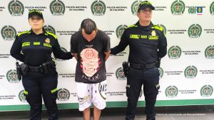 Wilmar Andrés Ruíz Cuenca está esposado con sus manos a la espalda, custodiado por dos uniformados de la Policía y de fondo un pendón de la institución.