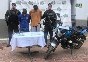 En la fotografía aparecen de pie, con los brazos atrás y la cabeza inclinada Fabián Andrés Urrego Balcázar y Jorge Alejandro Olmos Hernández. Uno, viste con camisa de manga corta y cuello, de color azul, con estampados blancos en el pecho; jean azul. El otro viste con camiseta de mangas cortas, de color naranja y una franja negro horizontal a la altura del pecho; así como pantalón gris. A los extremos hay uniformados de la Policía Nacional. Delante de ellos hay una mesa con mantel blanco y sobre ella hay un arma de fuego, así como dos letreros que dicen: ‘Arma de fuego’ y ‘Captura en flagrancia’. Al extremo izquierdo de la mesa hay una motocicleta. Detrás de los judicializados hay un pendón institucional de la Policía.