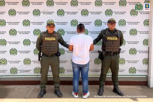 capturado de espaldas custodiado por dos policías, detrás banner de la Policía Nacional.