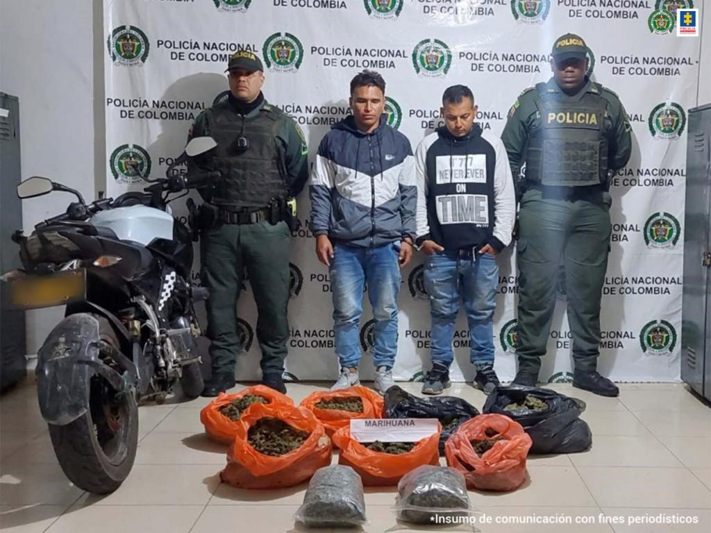 En la foto aparecen los dos motociclistas vestidos con chaqueta, buso y jean azul. A la derecha e izquierda de ellos hay dos uniformados de la Policía Nacional. Delante de ellos, a la derecha hay una motocicleta en frente a ellos, en el suelo, hay nueve bolsas de plástico, de colores naranja, negro y transparente y sobre ellas un letrero que dice marihuana. Detrás de ellos hay un pendón institucional de la Policía Nacional.