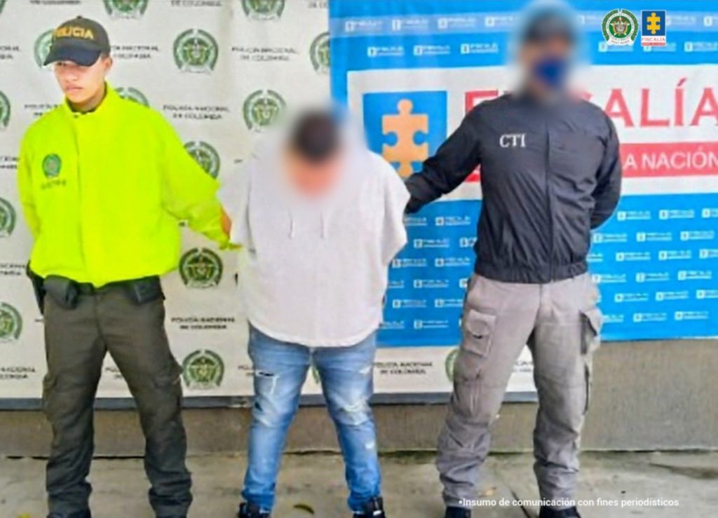 En la fotografía aparece el capturado junto a un funcionario del CTI y un uniformado de la Policía Nacional. En la parte superior está un banner de la Fiscalía General y otro banner de la Policía Nacional.