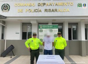 Se observa al capturado de frente, en el centro y su rostro cubierto con un efecto de difuminado. Atrás un pendón de Policía Nacional, adelante una mesa con el cartel "CAPTURA POR ORDEN JUDICIAL"