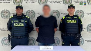 Se observa al presunto agresor de pie, con un efecto que le cubre el rostro y a cada lado suyo , un funcionario de la Policía Nacional. Atrás, un pendón del al Policía NAcional. Al frente, una mesa con el letrero violencia intrafamiliar. 