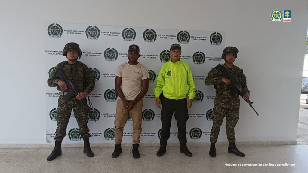 En la foto aparece el capturado de frente y custodiado por uniformados de la Policía  y Ejército Nacional. En la foto aparece de fondo un backing de la Policía Nacional.