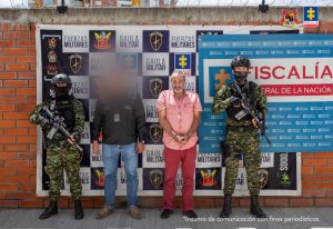 : El imputado se encuentra en custodia y tiene un pantalón rojo y camisa rosada, se encuentra en custodia por un servidor del Cuerpo Técnico de Investigación CTI, y dos uniformados del Gaula del Ejército Nacional. Detrás se observa un backing de la Fiscalía y Ejército.
