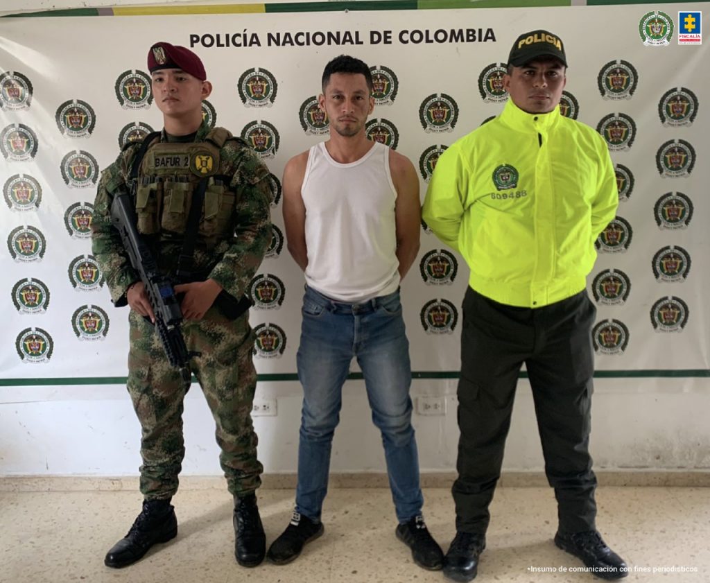 En la fotografía se observan tres personas, una de ellas el capturado, y las otras dos personas son uniformados de la Policía y el Ejército Nacional. En la parte posterior hay un backing con escudos de la Policía Nacional.