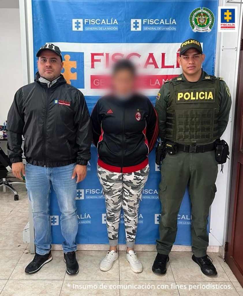 En la foto aparece la mujer judicializada, con los brazos atrás, vestida con buso negro y rayas rojas, legies tipo camuflado y zapatos tenis blancos. Al lado derecho de ella aparece un servidor del CTI de la Fiscalía y a su lado izquierdo uno de la Policía Nacional. Detrás de ellos aparece un pendón institucional de la Fiscalía.