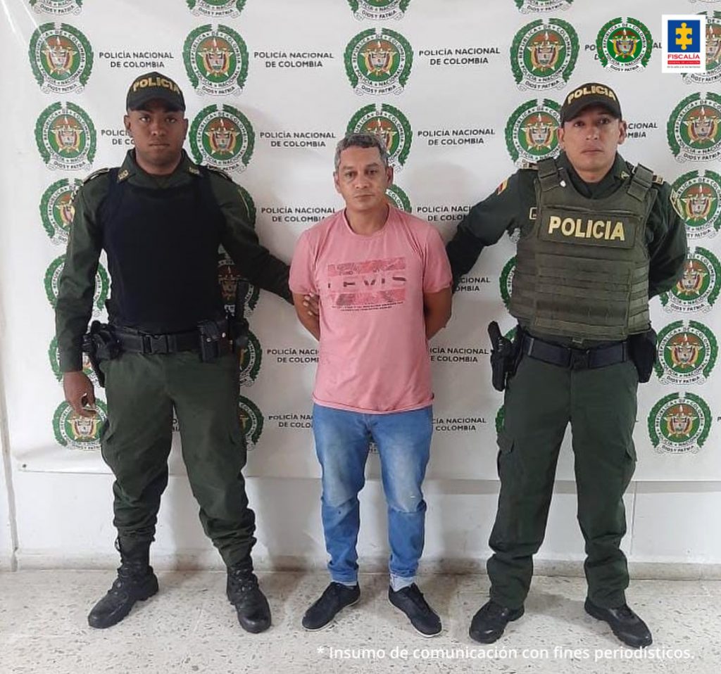 En la foto aparece el hoy imputado con los brazos atrás, vestido con camiseta rosada, con estapado a la altuera del pecho; jean azul y zapatos negros. A lado derecho e izquierdo aparecen dos representantes de la Policía Nacional. Atrás de ellos aparece un pendón institucional de la Policía Nacional.