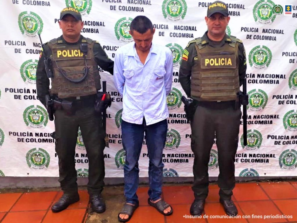 El capturado se encuentra esposado con sus manos a la espalda y custodiado por dos homnres de la policía nacional, atrás está en pendón institucional.