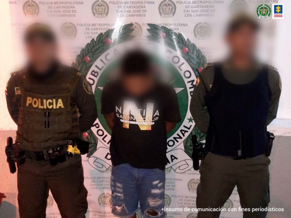 En la fotografía aparece el investigado custodiado por dos uniformados de la Policía Nacional. Detrás se observa el pendón de esa institución. Todos los de la imagen se encuentran con el rostro difuso.