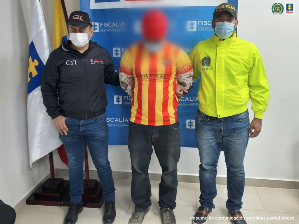 En la foto aparece el capturado con camiseta amarilla y roja, gorra roja con un uniformado del CTI y de la Policía Nacional que lo custodian, delante de un pendón de la Fiscalía General de la Nación.