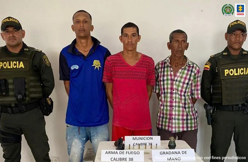 En la fotografía aparecen tres hombres, uno con camiseta azul, otro con camiseta roja y otro con camisa de cuadro de colores, custodiados por dos agentes de la Policía Nacional, con una mesa blanca donde aparecen las armas incautadas.