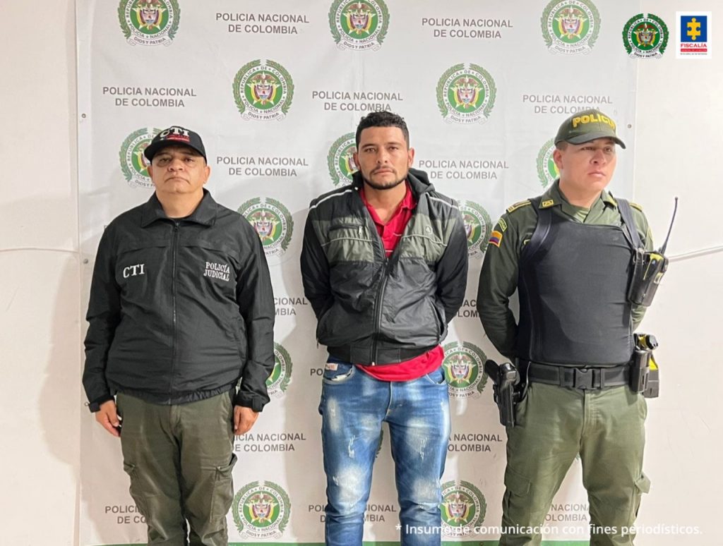 En la foto aparece el hoy imputado con los brazos atrás, vestido con camiseta roja, chaqueta y jean azul con manchas blancas. Al lado derecho aparece un servidor del CTI de la Fiscalía y la izquierda uno de la Policía Nacional. Detrás de ellos hay un pendón institucional de la Policía Nacional.