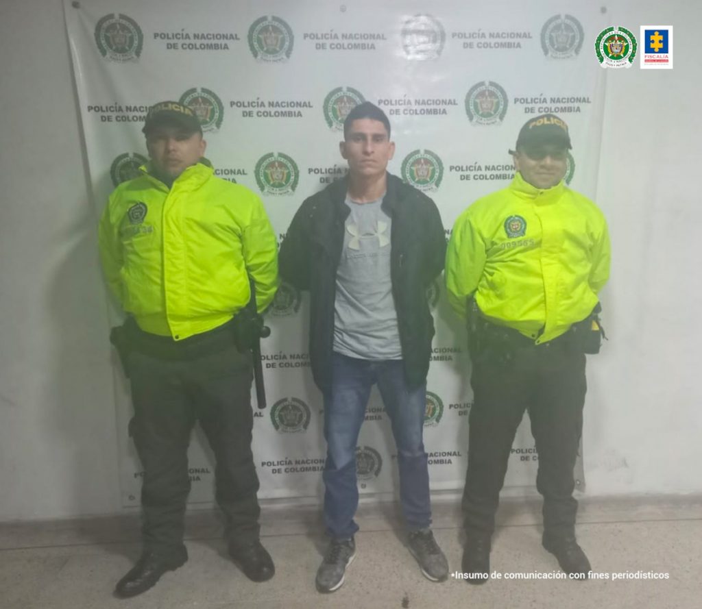En la fotografía se observa al capturado de frente a cada unop de sus lados un funcionario de la Policía Nacional. En el fondo se observa un pendón de Policía Nacional.