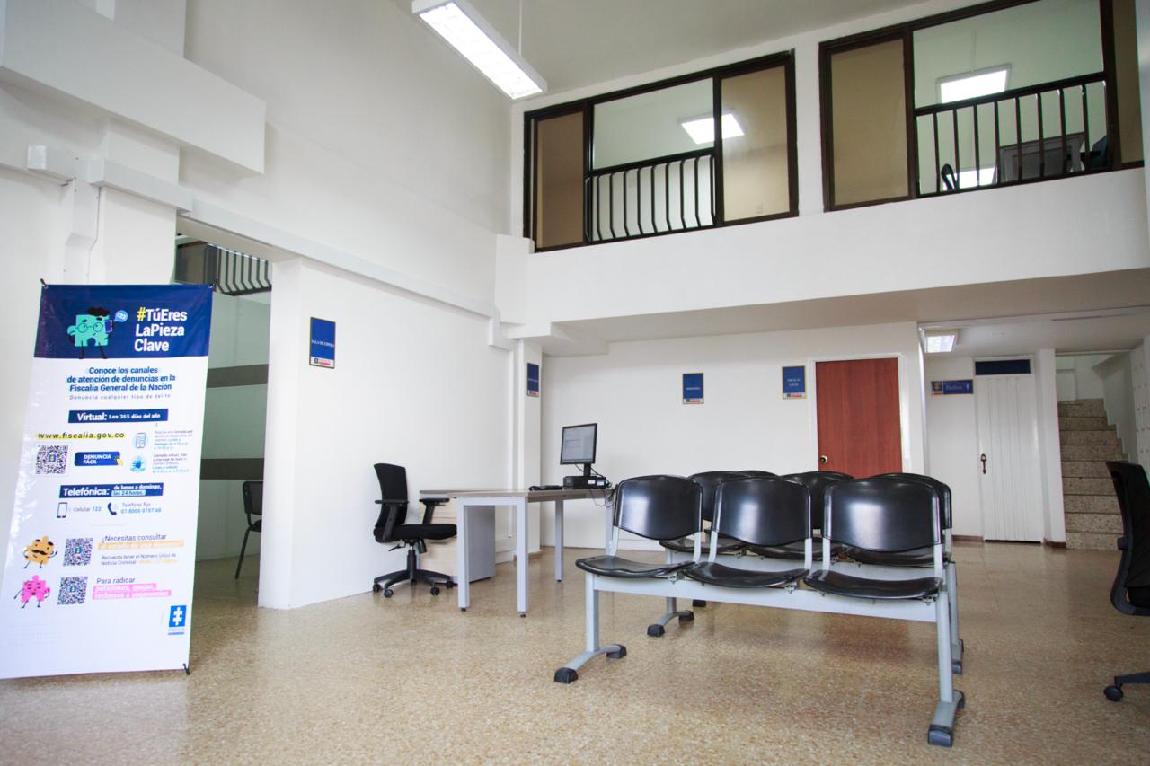Se ve el interior de una oficina de dos pisos. Tres sillas y un banner se ven en primer plano y algunos elementos de oficina, como un escritorio y un computador, se ven al fondo. 