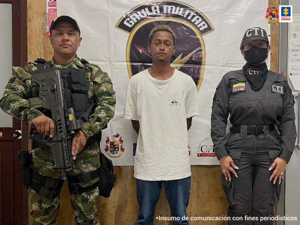 En la foto aparece el presunto implicado de extorsión tentada, con los brazos atrás, vestido con camiseta blanca y pantalón azul. A su lado derecho aparece un funcionario del Gaula Militar y a su izquierda una servidora del CTI de la Fiscalía. Atrás de ellos hay un pendón institucional del Gaula Militar con logos del Ejército y del CTI.