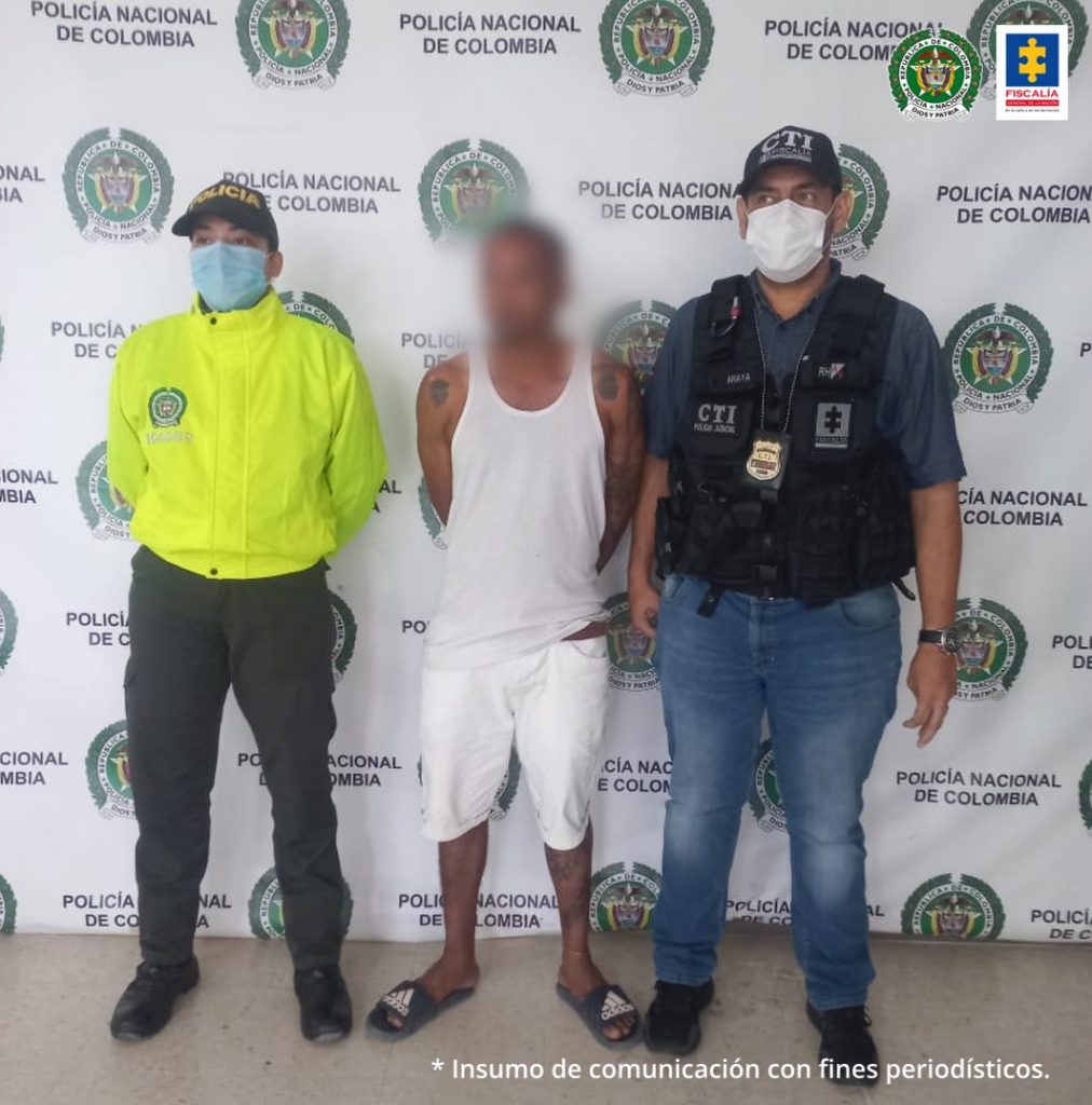 En la imagen aparece el procesado en camisilla blanca con bermuda blanca acompañada por un uniformado del CTI y un uniformado de la Policía Nacional, delante del pendón de la Policía Nacional.