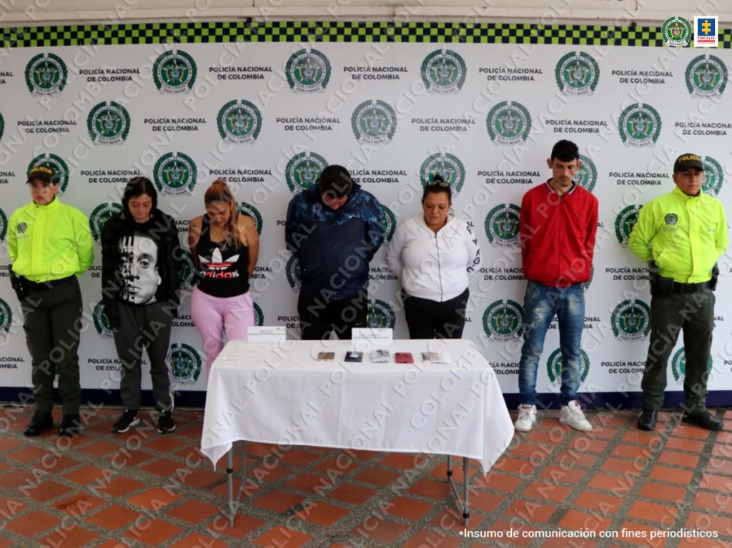 En la fotografía se observan cinco personas capturadas, tres mujeres y dos hombres, todos de frente. En cada extremo de la fila un funcionario de la Policía Nacional. Al fondo un pendón de Policía Nacional